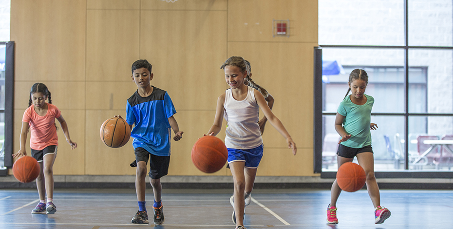 Four children are bouncing basketballs as they walk through a gymnasium