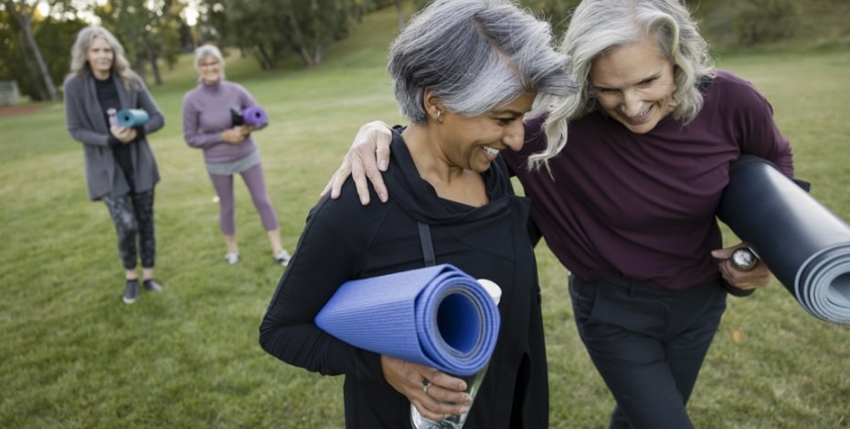 Two smiling ladies carrying yoga mats walking in the park.