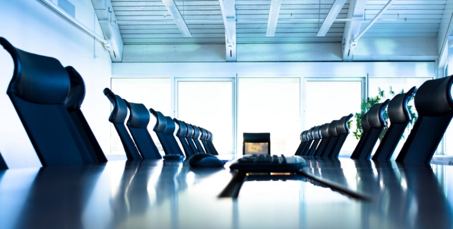 A selection of chairs in a boardroom all faced around a remote device sitting in the middle of a desk.