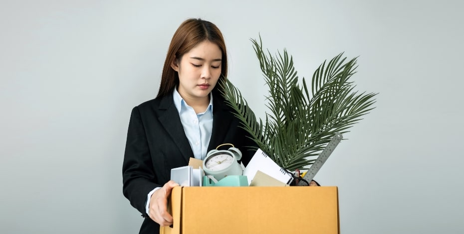 A sad woman is carrying a box of her possessions including a plant.