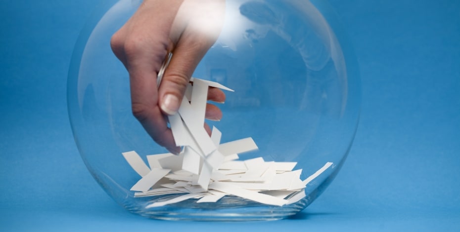 A human hand digs around pieces of paper inside a big glass bowl. 
