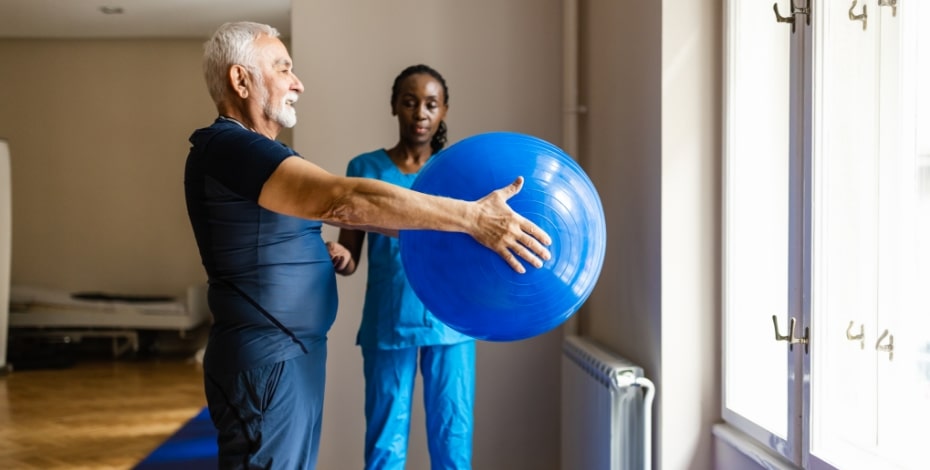 A physio supervises an older client holding a large inflatable ball.