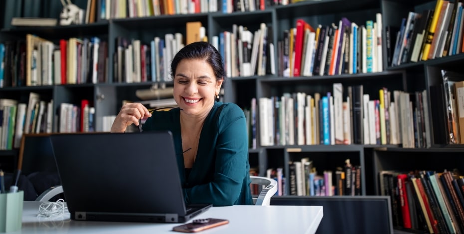 A woman in a green top sits at a computer in front of bookshelves.