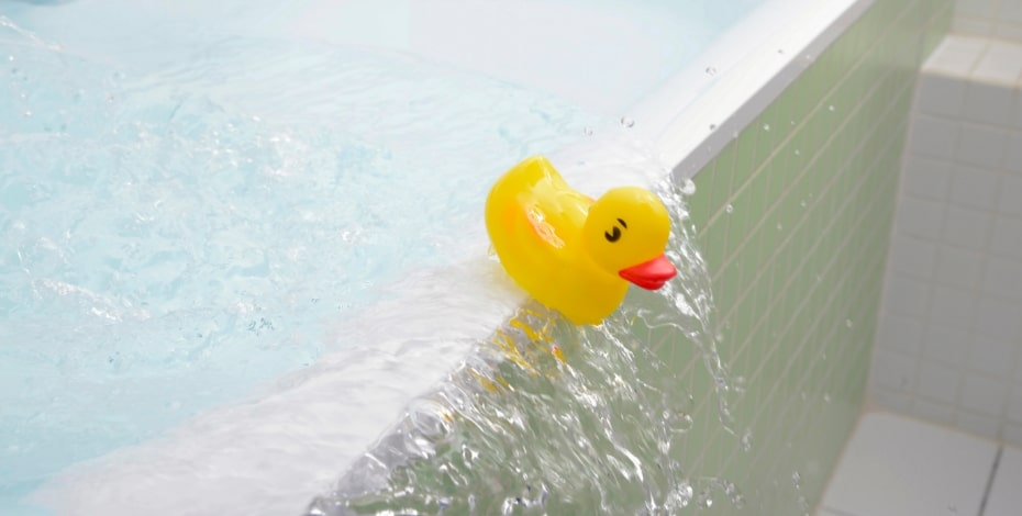 A little yellow rubber ducky spilling out of an overflowing bath tub.