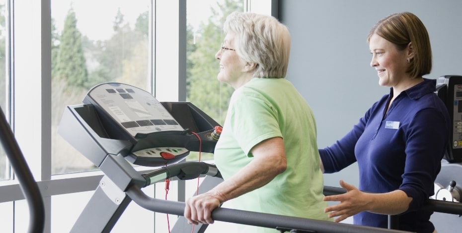 An older woman in a green t shirt and brown pants is on a treadmill in front of a window. A woman in a blue shirt and white pants is helping her. 