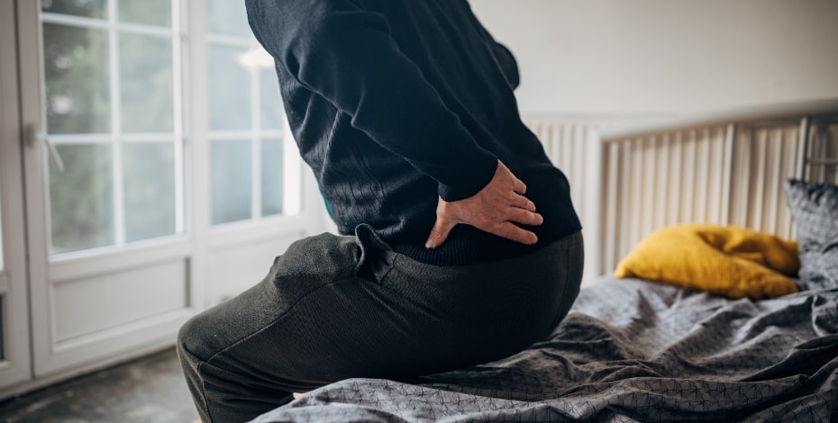 Image shows a person from the chest down holding their hip as they sit down on a bed. 