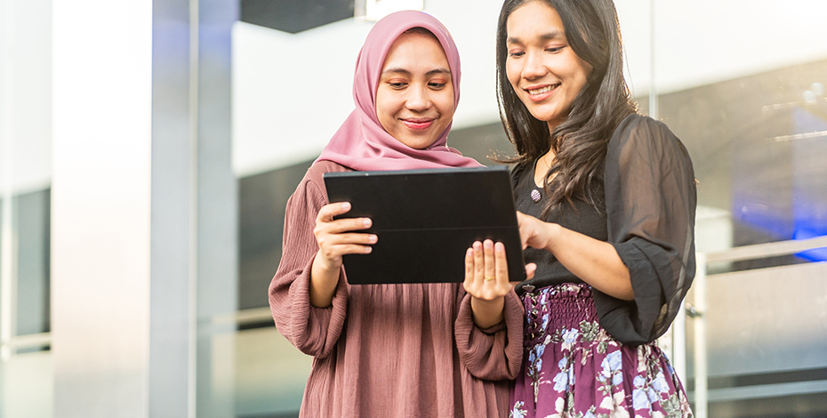 Two women stand around a computer tablet and are smiling while looking at the screen.