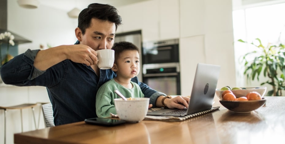 A man sips from a cup of coffee with a child sitting on his lap as they both look at an open laptop computer on the bench before them.