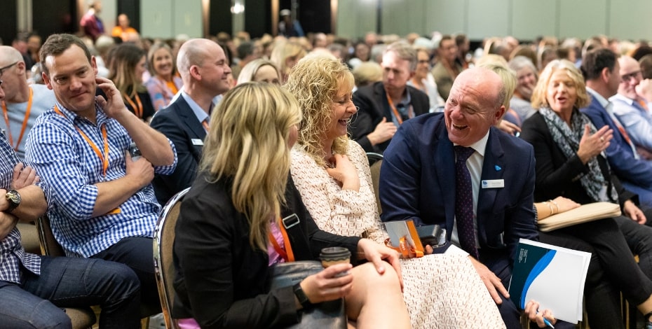 The image shows a group of people attending a conference. They are sitting in rows of chairs and talking to each other. 