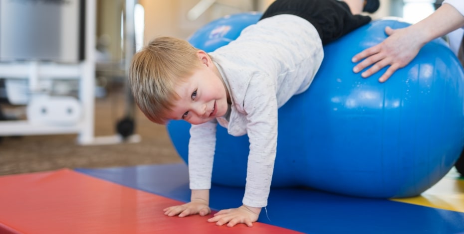 A child is held as he balances on a blue exercise ball. He is reaching down with his hands on the floor. 