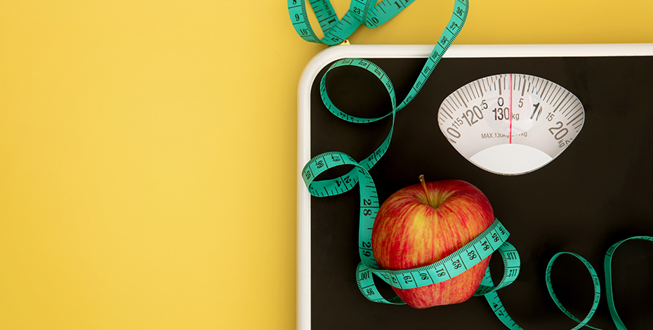 A set of bathroom scales against a yellow background, with an apple and a tape measure