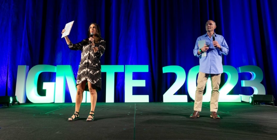 A man and a woman stand on a stage in front of the words IGNITE 2023. There is a blue curtain behind them. They are holding notes and microphones. 