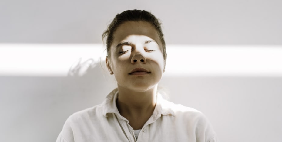 A woman stands against a white wall with the reflection of the sun across her face, symbolising a release from pain.