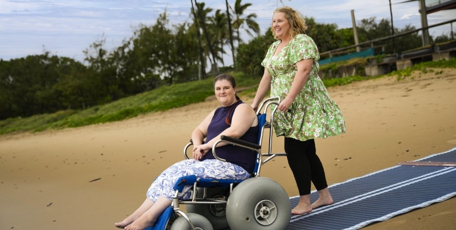 a blonde woman in a green dress and black leggings is pushing another woman in a wheelchair fitted with sand wheelsdown to the beach along a mat.  