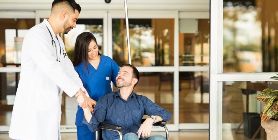 A clinician wearing a white lab coat is talking to a man in a wheelchair. A nurse in blue scrubs is pushing the chair. 