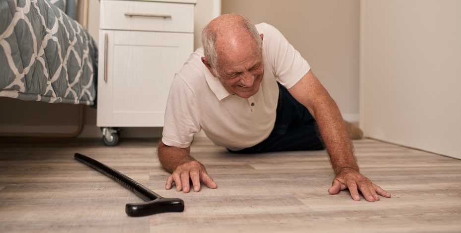 An elderly man has fallen over next to his bed. A walking stick is on the floor next to him. 