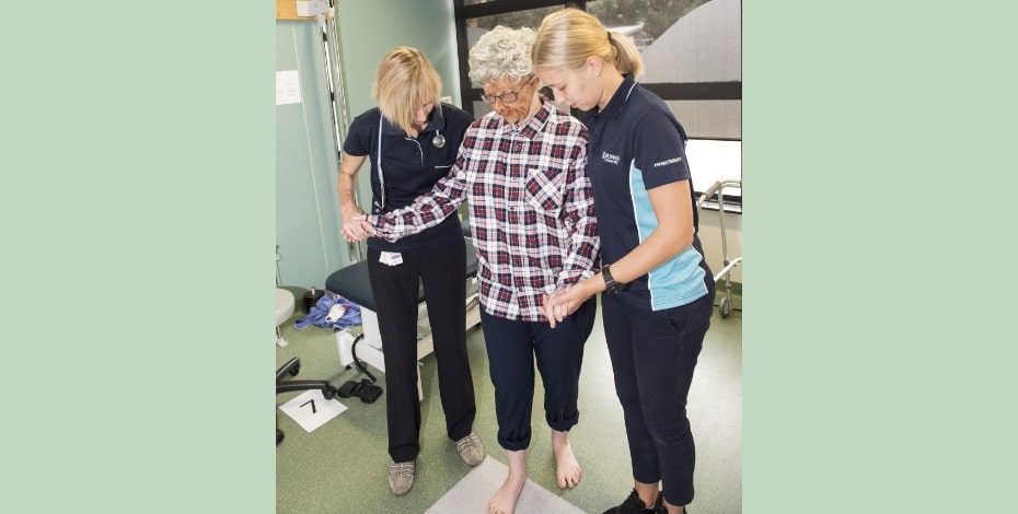 student physiotherapists assist a patient (played by another student). The patient is wearing clothes and a mask to make them appear elderly. 
