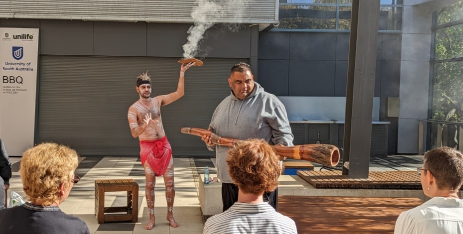 An Aboriginal man shows his audience a digeridoo, while another Aboriginal man wearing a red loin cloth holds up a shield with smoking leaves. They are conducting a smoking ceremony