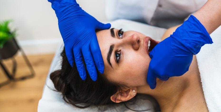 A woman is lying on an examination table. A person is manipulating her jaw. They are wearing blue gloves on their hands. 