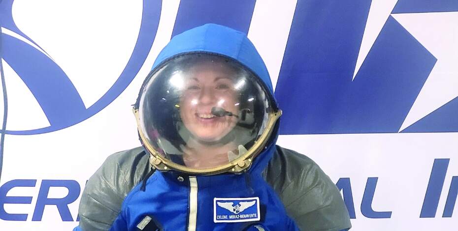 A young woman is wearing a blue spacesuit. She is smiling through the faceplate. 