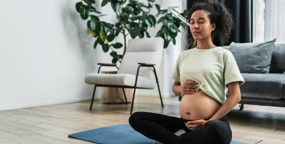 A pregnant woman is sitting cross-legged on the floor. Her green t-shirt has been pulled up and she is cradling her belly.. 