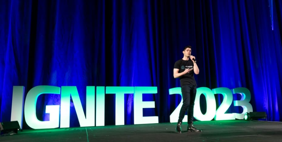 A man wearing black t shirt and black pants is giving a speech. He is standing on a stage in front of the words IGNITE 2023. 