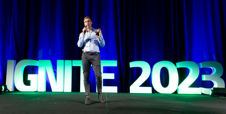 A man is standing on a stage in front of the words IGNITE 2023 and talking into a microphone