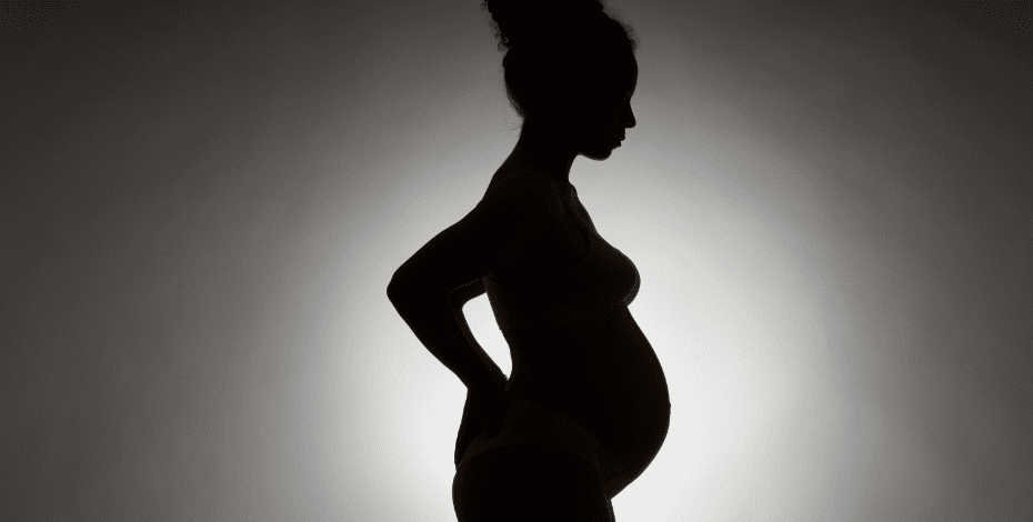 A pregnant woman stands in darkness, with a white light behind her