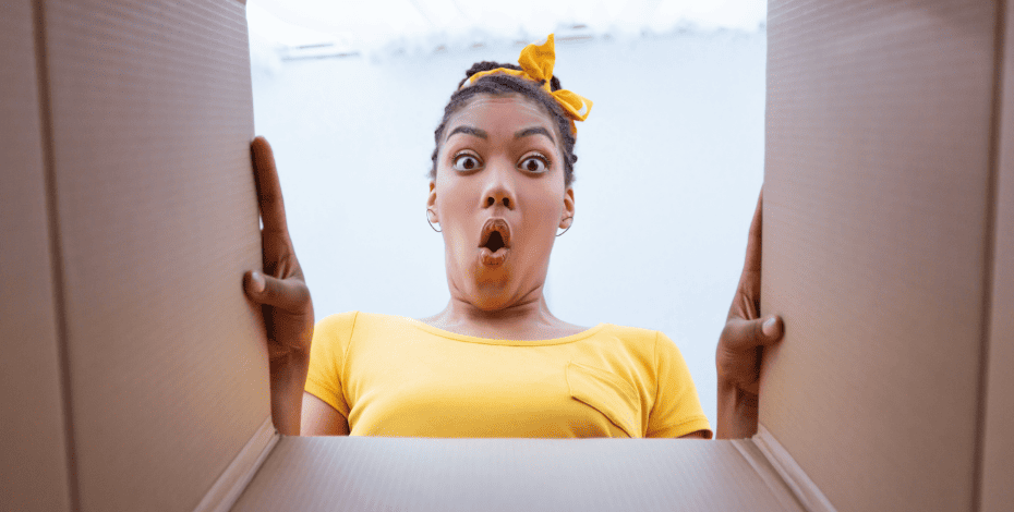 A woman with brown skin wearing a bright yellow t shirt is looking through the opening of a cardboard box. 