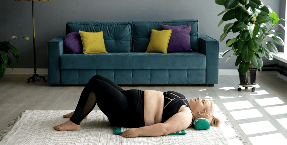 A larger woman wearing exercise clothes is lying on a mat on the floor with a neck pillow supporting her head. 