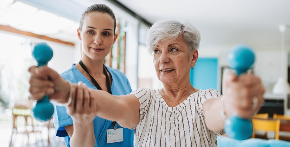 A physiotherapist assists an older woman to lift weights in a clinical setting.