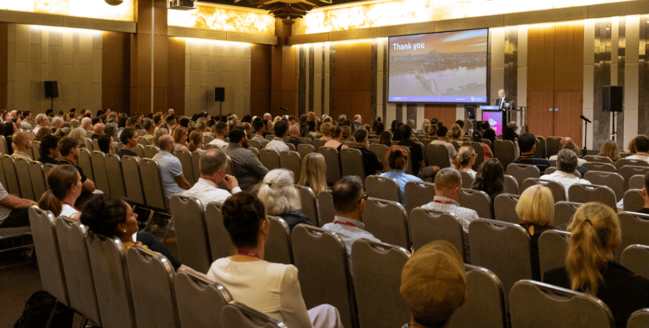 The image shows people sitting in a conference session listening to someone speaking at a podium up the front. The words Thank You are shown on the screen. 