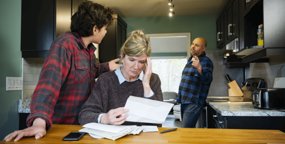 The image shows a family in a kitchen. A women is sitting at a table reading paperwork. Behind her, her male partner and her child are arguring. 