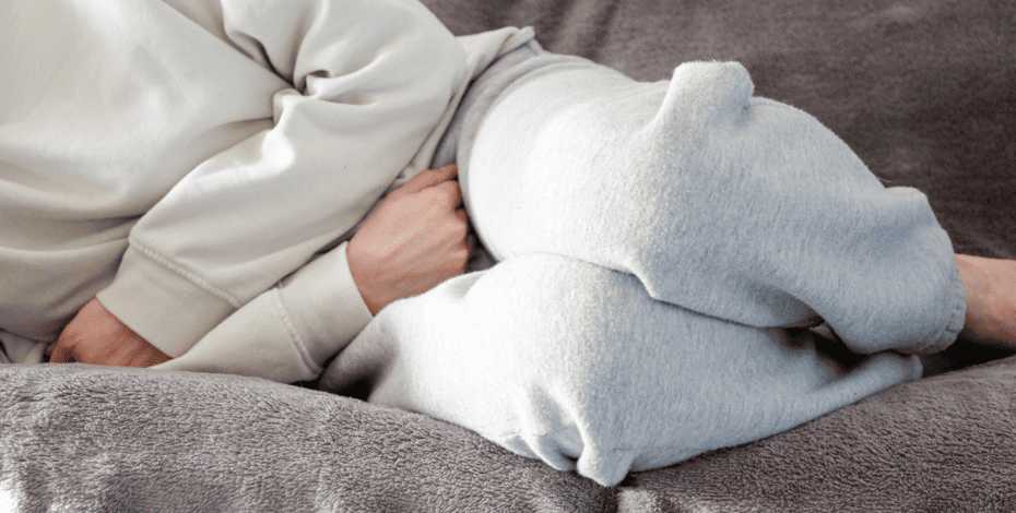 The image is of a person in pale grey sweats lying on a couch with their arms holding their abdomen. The person's head is not in the image. 
