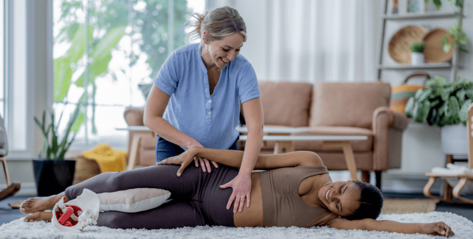 The image shows a pregnant woman lying on a rug on her side. A physiotherapist has her hands on the woman's hip and belly. 