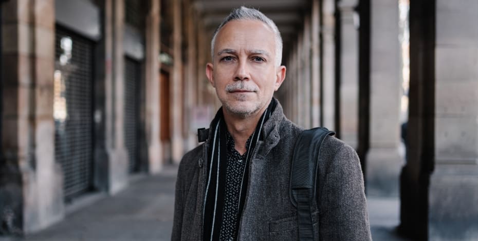 The image is of an older man standing outside on a walkway by a building. 