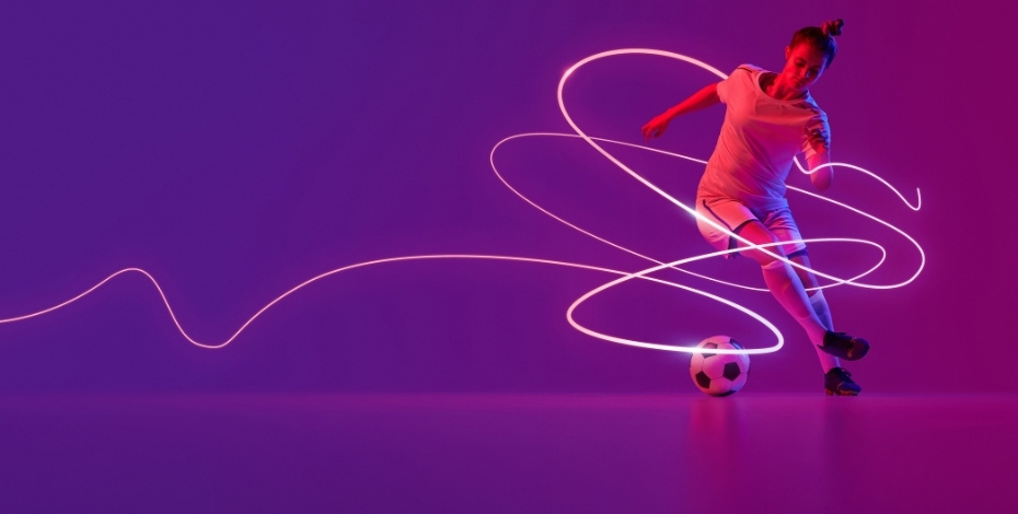The image is of a female soccer player kicking a soccer ball, on a purple background. A wavy white line wraps around the soccer player to provide dynamic movement. 