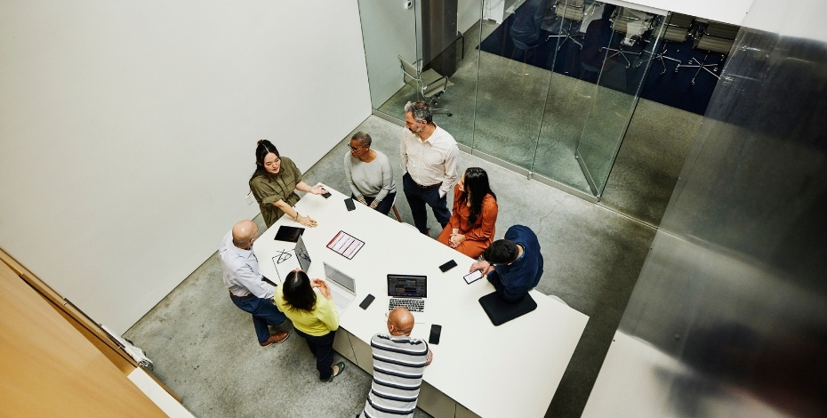 The image looks down from above at a group of workers in discussion around a table. There are several files and laptops on the table. 