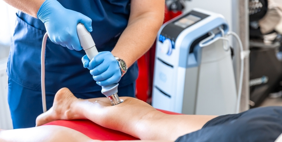 A man is lying on an examination table receiving shockwave therapy to his calf from a physiotherapist