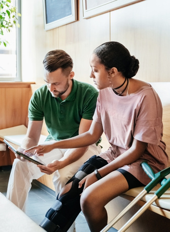 A woman with her leg in a brace and crutches is showing the man sitting next to her something on a tablet screen. 