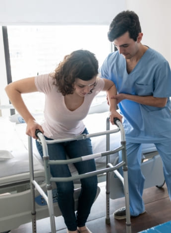 The image is of a male physiotherapist assisting a female patient to stand up using a mobility frame. 