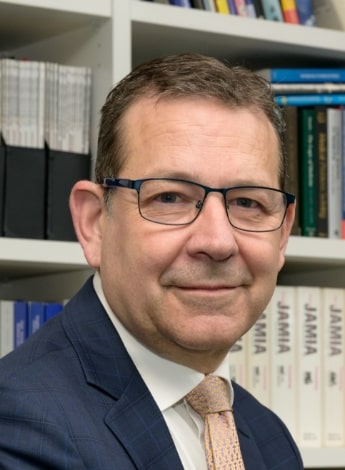 The image is of a man wearing glasses in a suit and tie. He is in front of a bookshelf