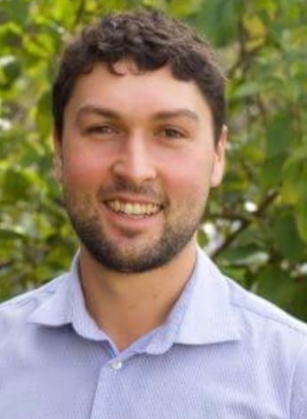 A man with dark brown curly hair and a short beard is standing in front of greenery.