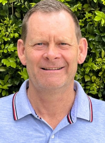 An older man is standing in front of greenery. He is wearing a blue collared shirt with a red trim. 