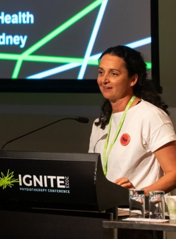 A woman with long dark hair is talking at a podium with the words Ignite 23 on it. Part of her presentation slide can be seen behind her. 