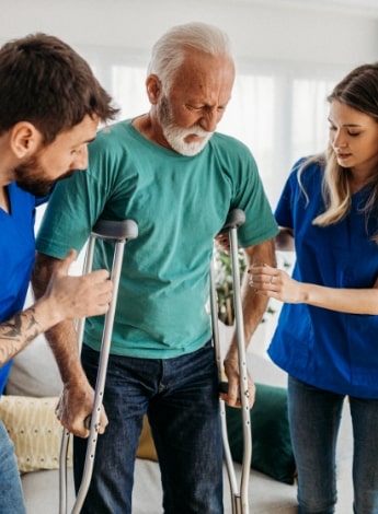 An older man on crutches is assisted by two carers.