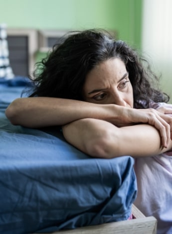 A sad-looking woman leans on the corner of a bed.