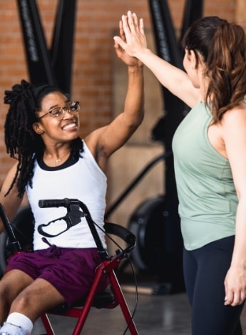 A woman in a wheelchair high-fives a standing woman. Both in gym clothes.