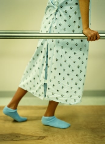 A person in a hospital gown and socks walks holding onto parallel bars. 