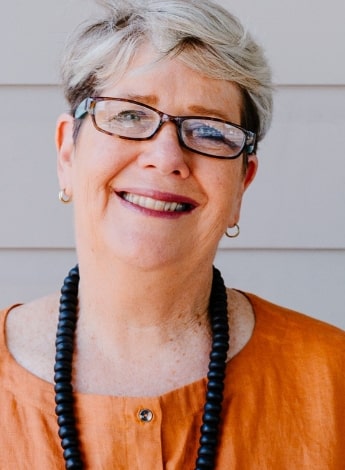 ""A woman with short gray hair and glasses is smiling at the camera. She is wearing an orange shirt and a chunky black necklace.